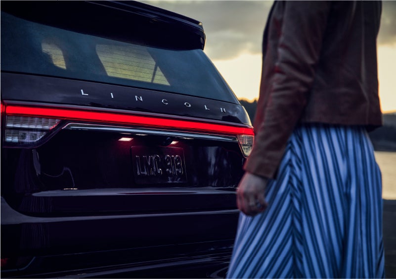 A person is shown near the rear of a 2023 Lincoln Aviator® SUV as the Lincoln Embrace illuminates the rear lights | Lexington Park Ford Lincoln in California MD