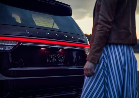 A person is shown near the rear of a 2024 Lincoln Aviator® SUV as the Lincoln Embrace illuminates the rear lights | Lexington Park Ford Lincoln in California MD