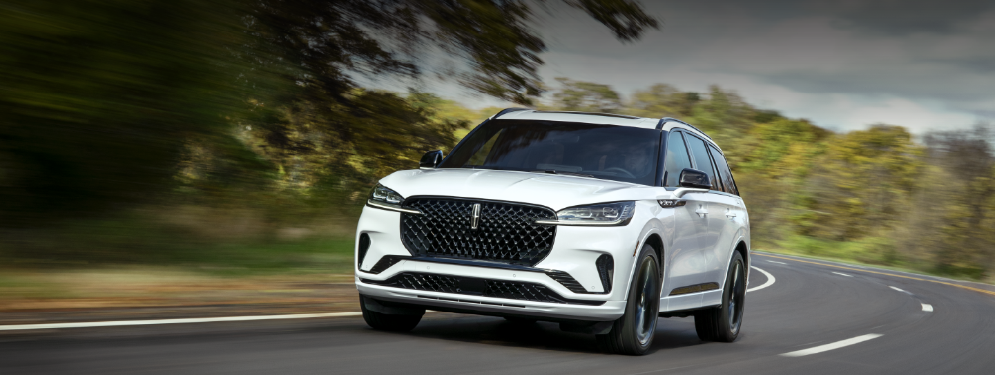 A white 2025 Lincoln Aviator® SUV is being driven down a road lined with trees. | Lexington Park Ford Lincoln in California MD
